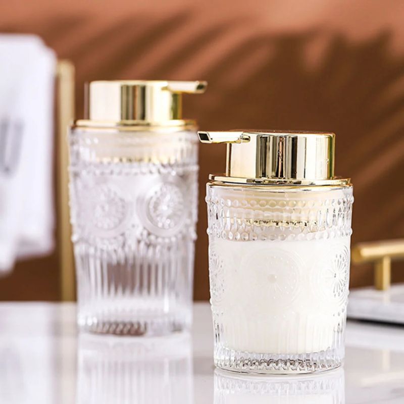 Two clear glass soap dispensers with ornate patterns and gold pumps, standing on a light marble surface. One dispenser is filled with white liquid.