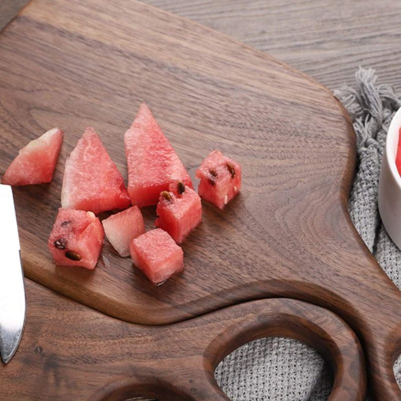 A close-up shot shows a wooden cutting board with a distinctive handle featuring a circular cutout. Several chunks and slices of fresh watermelon, with their vibrant pink flesh and black seeds, are scattered across the board. A portion of a silver knife is visible on the left edge of the board. The cutting board rests on a light gray, textured surface, possibly a placemat or tablecloth. The lighting is bright, highlighting the textures of the wood and the juicy appearance of the watermelon.