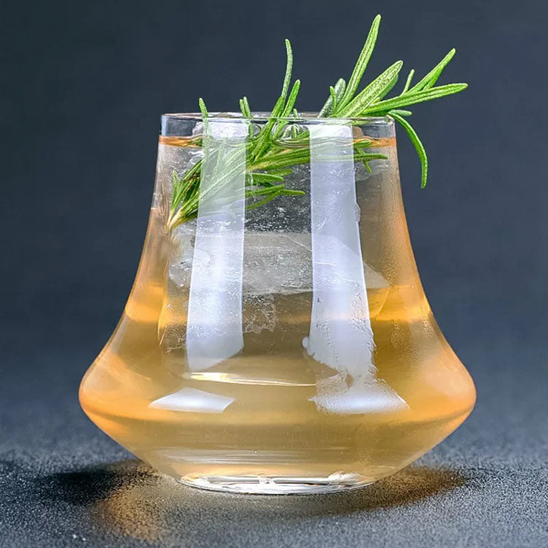 A uniquely shaped whiskey glass filled with a golden cocktail, garnished with fresh rosemary and ice, against a dark textured background.