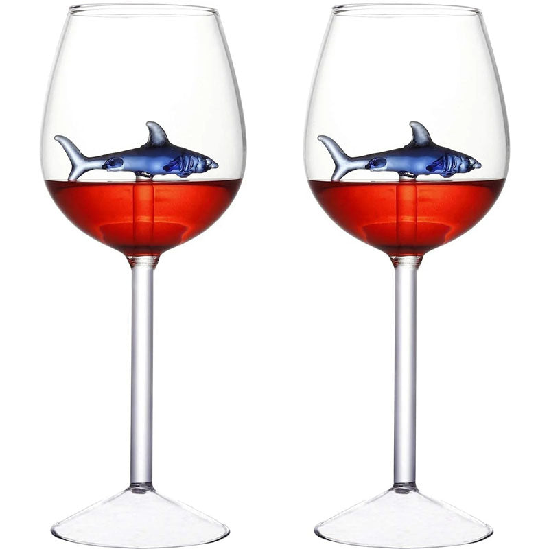 A pair of wine glasses filled with red liquid, each containing a small, blue glass shark figurine.