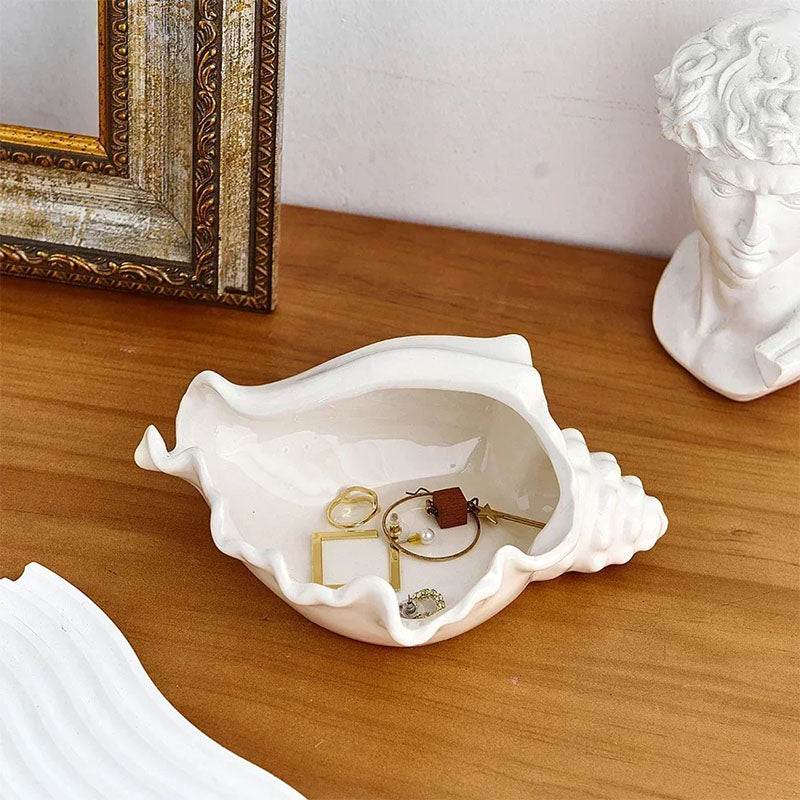 A white ceramic seashell-shaped jewelry dish on a wooden surface, holding gold rings and earrings, with a decorative frame and a classical-style bust nearby.