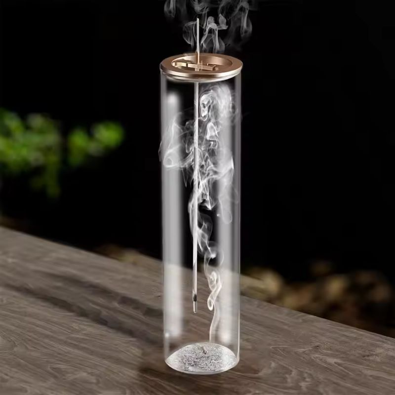 This image features a modern glass incense holder with a gold-colored lid, placed on a wooden surface. Inside the transparent cylindrical container, an incense stick is burning, releasing delicate swirls of smoke that rise gracefully. The base of the holder contains a layer of ash. The background is dark and blurred, with hints of green foliage, creating a serene and elegant atmosphere.