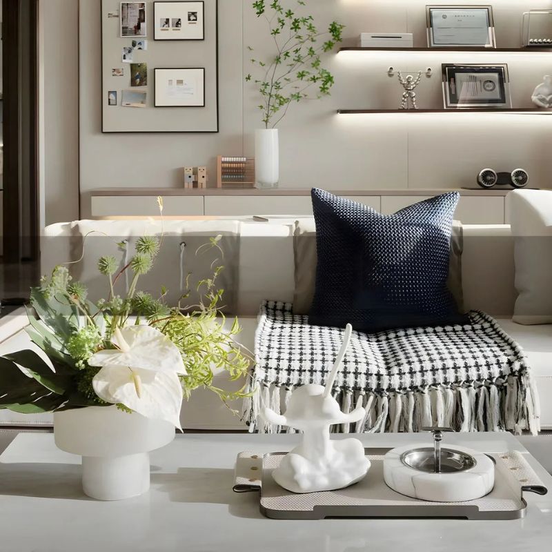 A modern living room with a neutral-toned sofa adorned with a navy blue textured pillow and a black-and-white checkered throw blanket. A coffee table in the foreground features a white ceramic vase with a lush floral arrangement, a sculptural white figurine, and a decorative tray. In the background, illuminated wall shelves display framed certificates, books, and artistic decor, while a framed photo collage and a tall white vase with greenery add a personal touch.