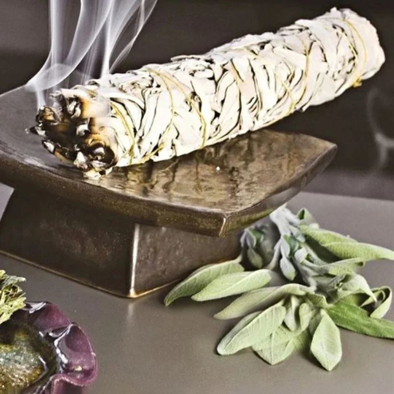 A close-up shot of a thick bundle of white sage, partially burned and smoking, resting on a small, square, dark-colored stand with a slightly reflective surface. The sage bundle is tied with yellow string and shows signs of burning on one end. Wisps of white smoke rise from the burning sage. In the lower right corner, sprigs of fresh green sage leaves are visible, and in the lower left corner, a partial view of a purple object is seen. The background is a blurred, neutral tone.