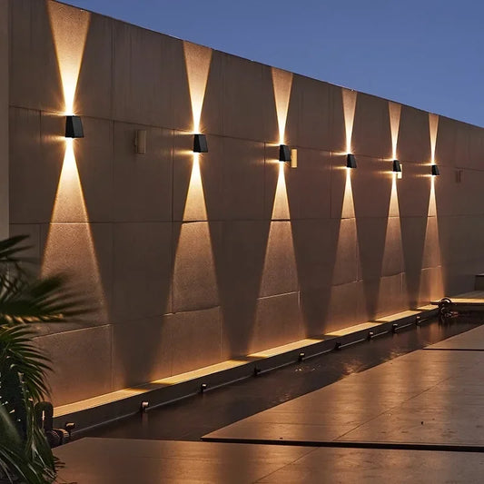 Six small, dark, rectangular lights are evenly spaced and mounted on a textured wall, each casting a warm white triangular beam both upwards and downwards at dusk or night. A reflecting pool runs along the base of the wall.