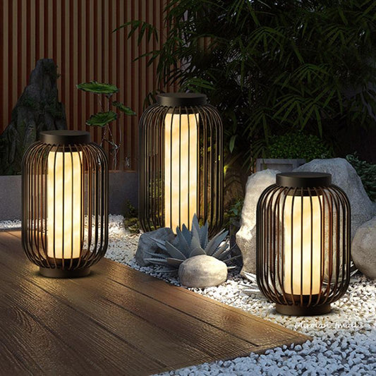 Three modern cylindrical outdoor lanterns with dark frames and warm internal lighting illuminate a garden area at night. The lanterns are placed on a wooden deck and white gravel, surrounded by rocks and plants.