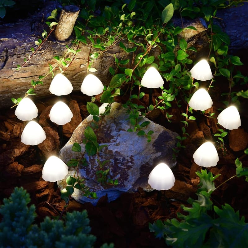 Numerous small, mushroom-shaped solar lights with translucent white caps emit a bright white glow in a garden at night. The lights are on thin dark wires and are scattered among rocks, vines, and dark green foliage.