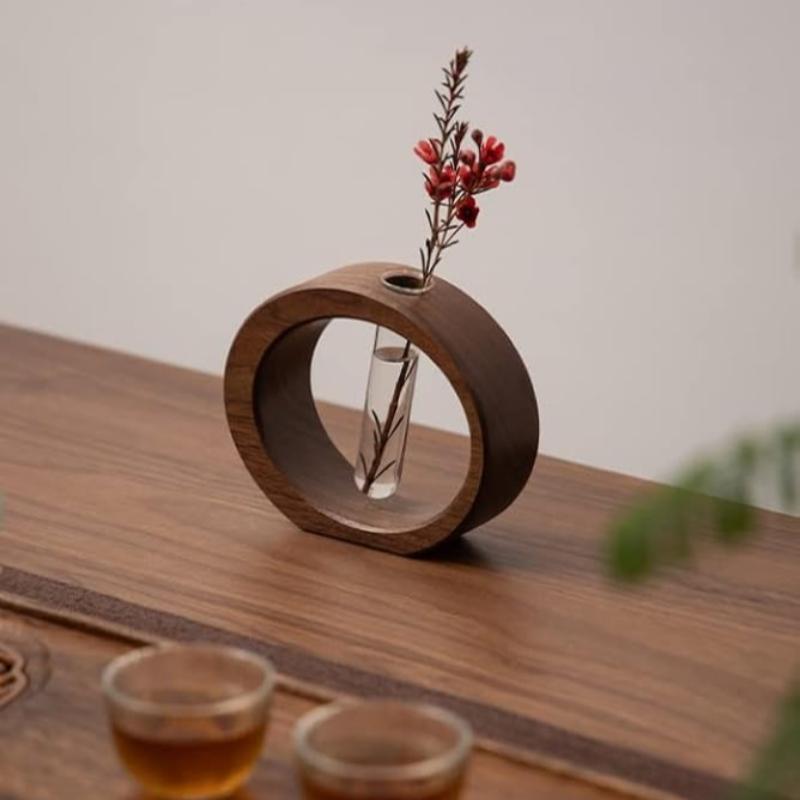 A dark wooden circular vase holds a clear glass test tube with a delicate stem and small red flowers. It rests on a carved wooden surface, with two small glasses filled with a light brown liquid partially visible in the foreground.