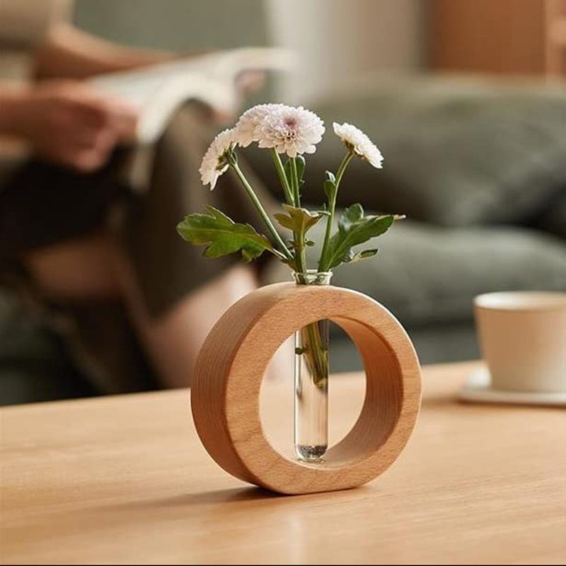 A light wooden circular vase holds a clear glass test tube with several small white chrysanthemums and green leaves. It sits on a light wooden table.