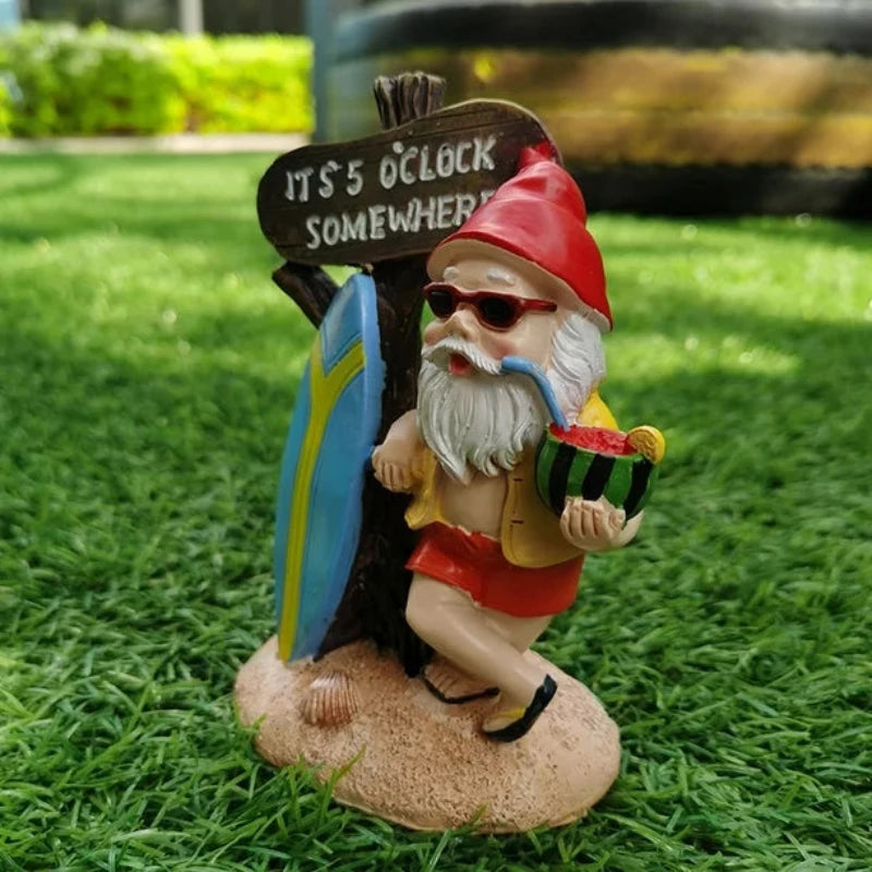 A whimsical garden gnome figurine with a long white beard and sunglasses, wearing a red pointed hat, a yellow open shirt, and red shorts with black sandals, stands on a sandy base with seashells. He holds a slice of watermelon in one hand and a pipe with a blue mouthpiece in the other. Behind him is a rustic wooden sign that reads "IT'S 5 O'CLOCK SOMEWHERE," leaning against a blue and yellow surfboard. The background is bright green artificial grass.