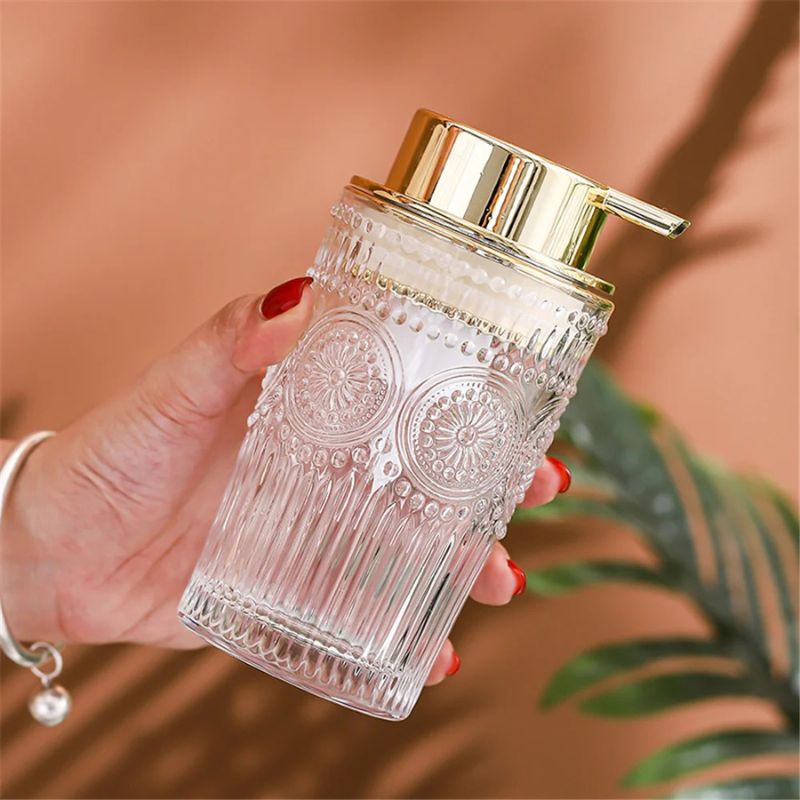 A hand with red nails holding a clear glass soap dispenser with ornate patterns and a gold pump. A bracelet is visible on the wrist.