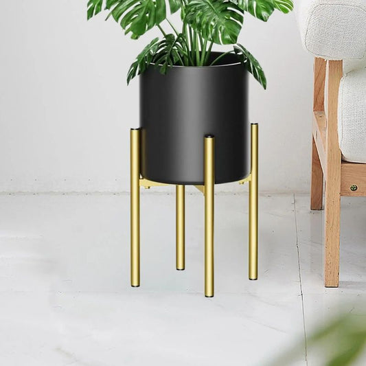 A modern indoor planter featuring a sleek black cylindrical pot elevated by a gold metal stand with four slim legs. The planter holds a vibrant green monstera plant, adding a touch of nature to the contemporary setting. Positioned next to a wooden-framed, white-upholstered sofa, the planter complements the minimalist and stylish interior decor.