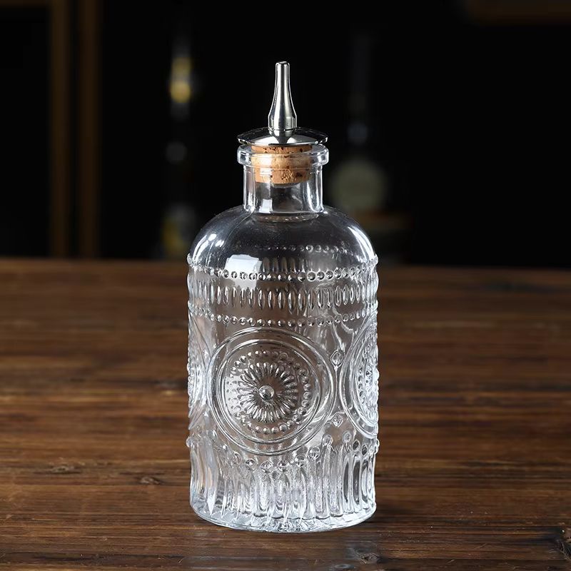 Clear glass bitters bottle with intricate embossed patterns, a cork stopper, and a metal pour spout, set on a wooden surface.