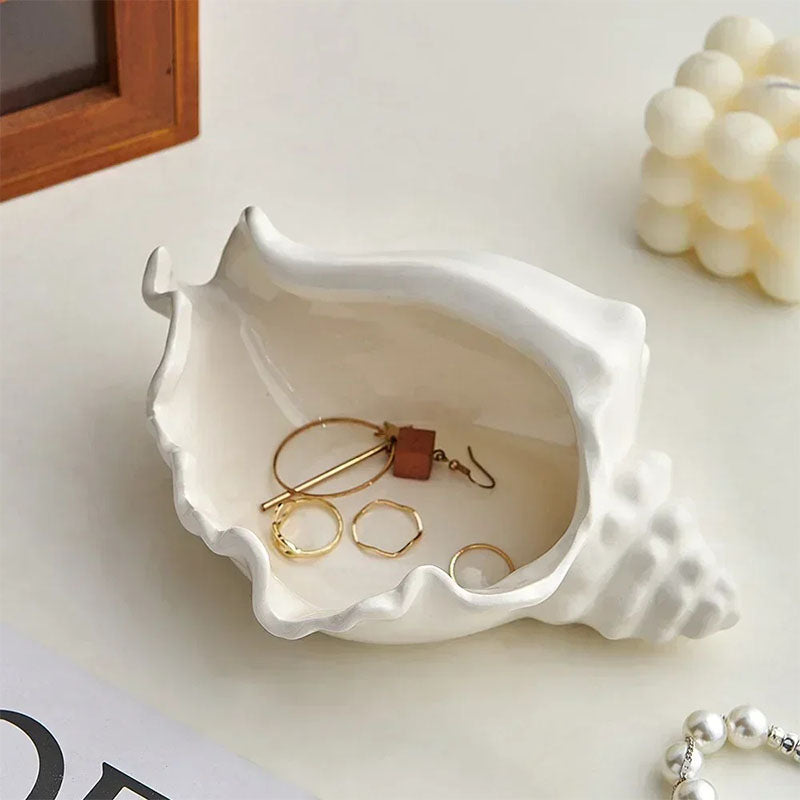 A white ceramic seashell-shaped jewelry dish on a white surface, holding gold rings and earrings, with a pearl bracelet, a wooden frame, and a decorative candle nearby.