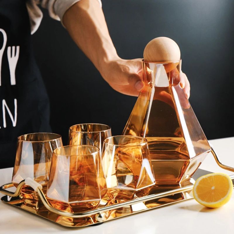 Amber glass pitcher and four faceted glasses on a gold tray, with a hand reaching in. A lemon slice is on the tray, and the letter "N" is visible on a dark background.