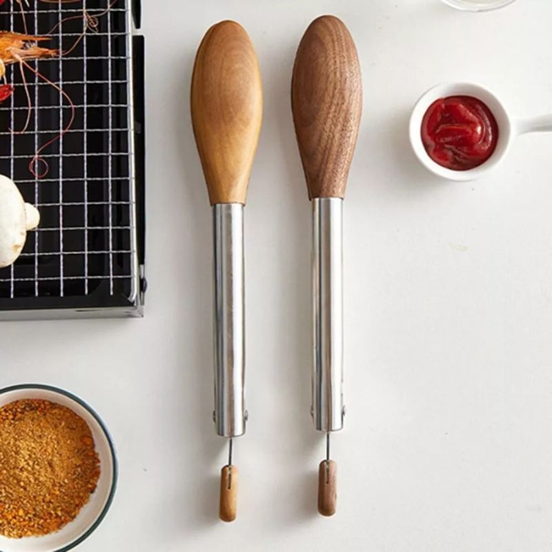 Two stainless steel tongs with wooden handles, one light wood and one dark wood, laid vertically on a white surface. A small grill, a bowl of spices, and a small dish of ketchup are also visible.