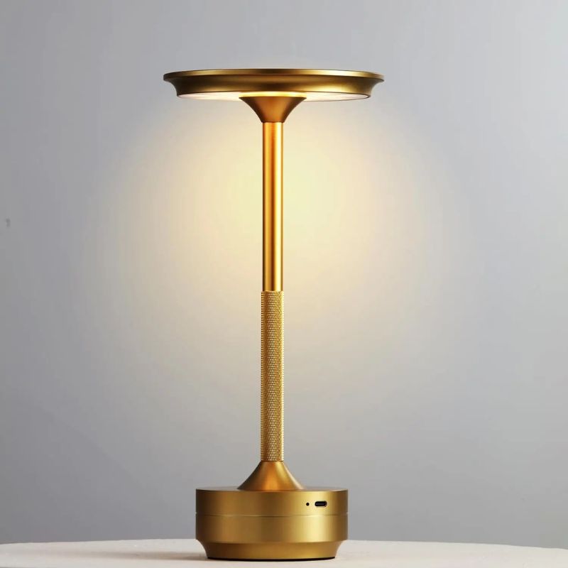 Sleek gold LED table lamp with a disc-shaped light head and a textured stem, standing on a smooth surface against a neutral background. The lamp emits a warm, ambient glow.
