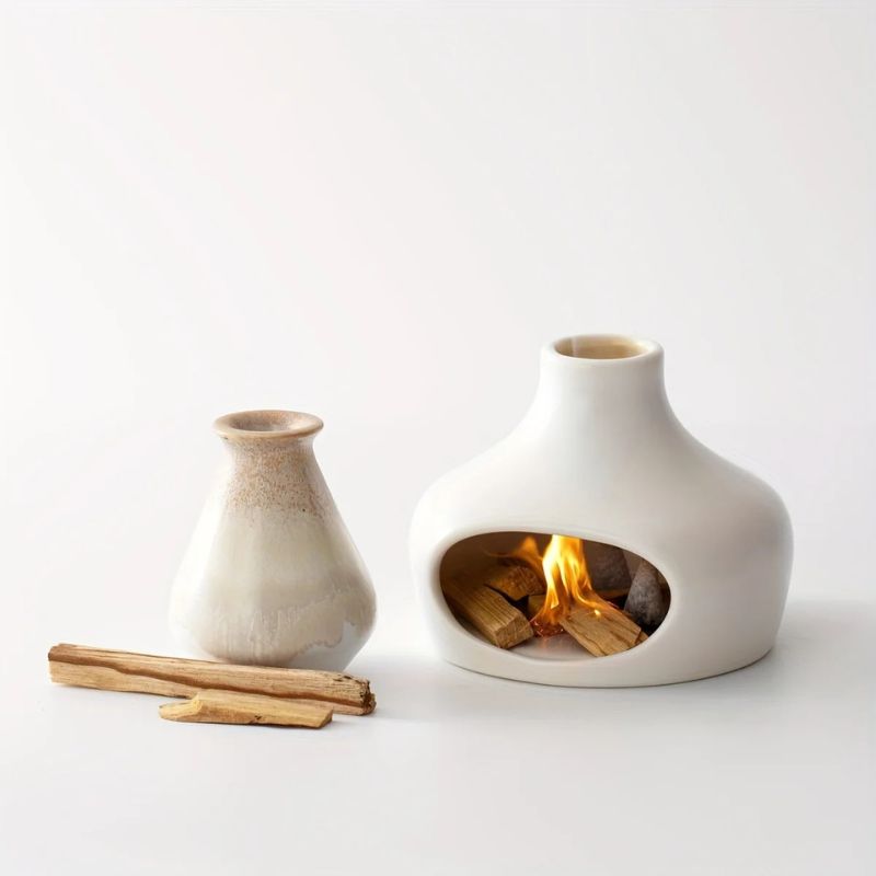 A white ceramic incense burner with a fire inside, emitting smoke through the top opening, placed next to a smaller glazed ceramic vase. A few wooden incense sticks are laid on the white surface beside them.