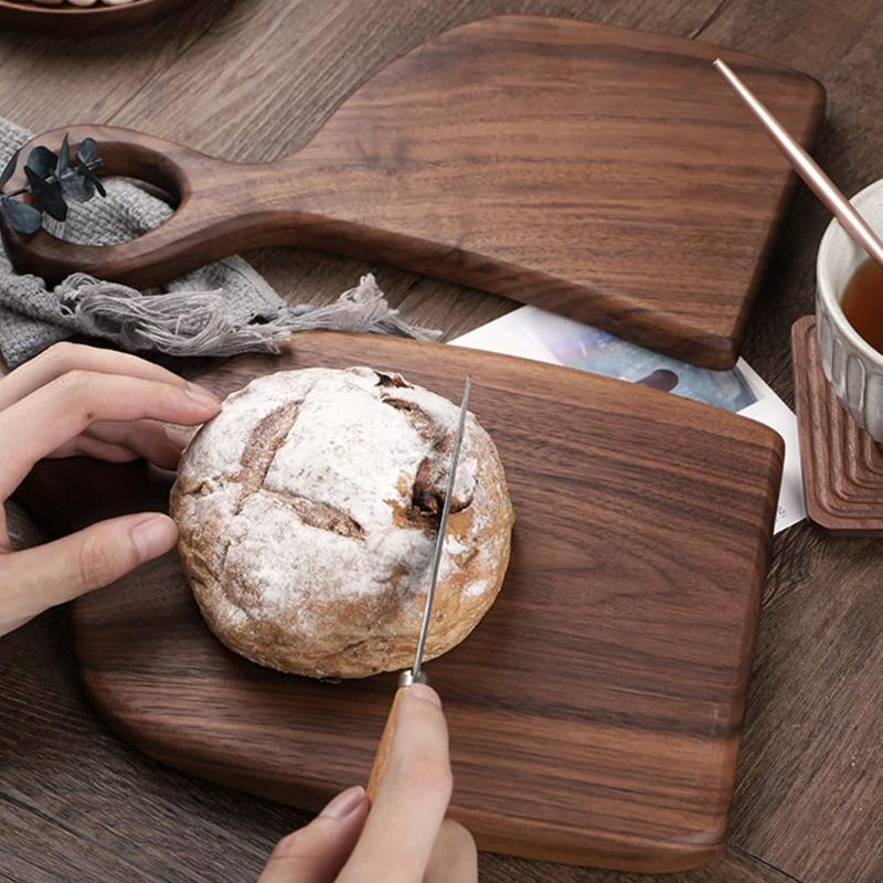 Two wooden cutting boards with circular handles are on a dark wooden surface. A person's hands are slicing a round loaf of bread on the smaller board. A mug with a light-colored beverage sits on a wooden coaster near the larger board. A gray cloth and a partially visible book are also present.