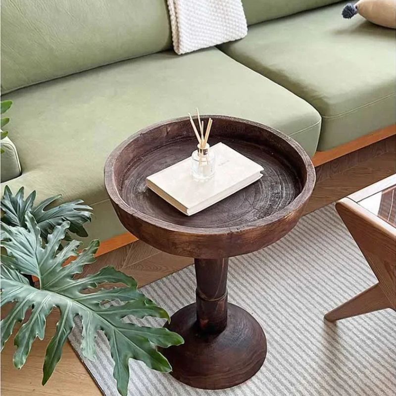 A rustic, dark wood, round side table (Momo Wabi-Sabi Log Coffee Table) with a shallow tray top, holding a book and a reed diffuser, positioned next to a green sofa and a striped rug.