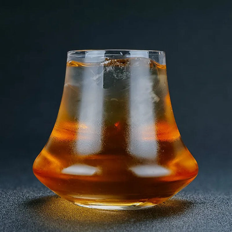 A stylish whiskey glass with an elegant, flared shape, filled with amber liquid and ice, set against a dark background.
