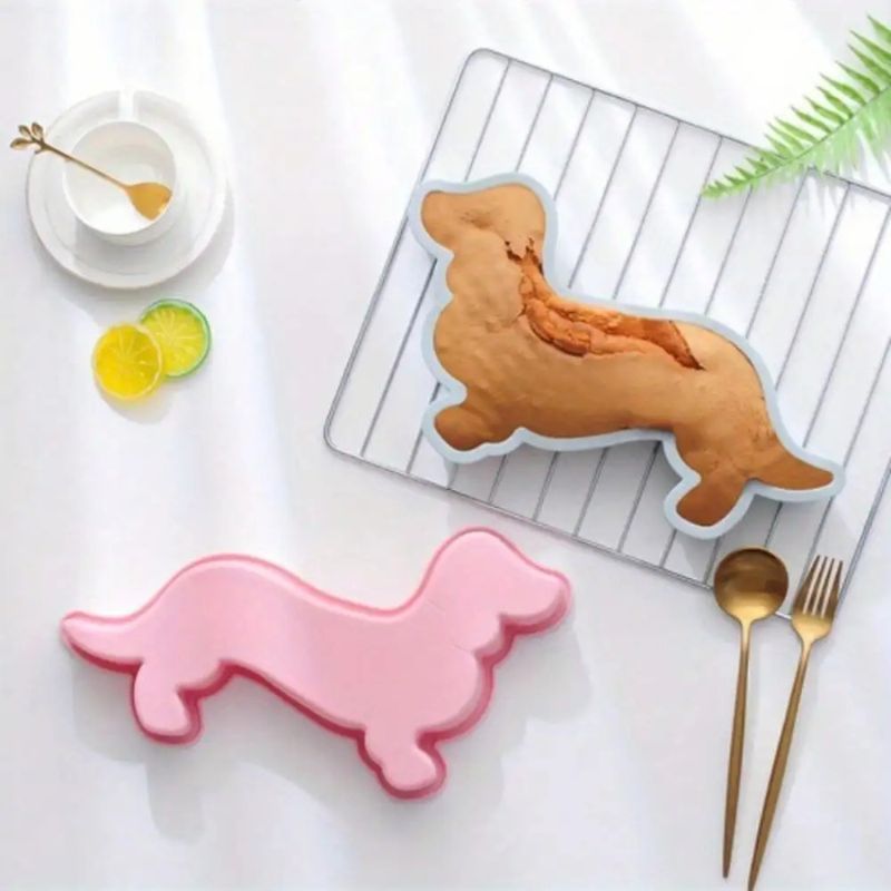 Overhead shot of a baked dachshund in a light blue mold on a wire rack, a pink dachshund mold, and gold utensils on a white surface with shadows. A white cup and saucer with citrus slices are in the upper left.