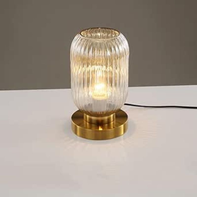 A lit, ribbed amber glass table lamp with a gold base sits on a white surface against a plain gray background. This could be a TaleTre Modern Glass Table Lamp.