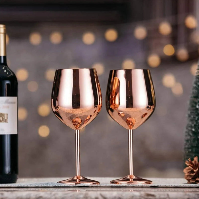 Two elegant, copper-toned stemmed wine glasses stand on a textured surface next to a dark wine bottle. The background is softly blurred with warm, out-of-focus lights, suggesting a festive setting. A small pinecone is visible on the right.