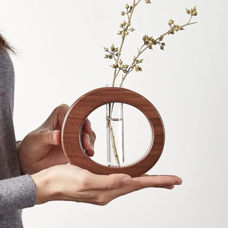 A circular wooden vase holds a clear glass test tube with a dried plant stem. The vase is being held by a person.