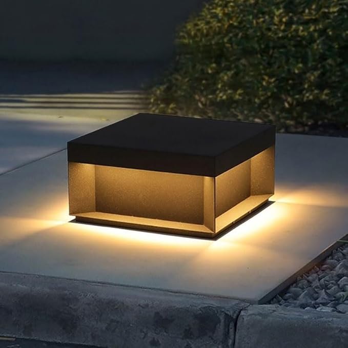 A modern, square outdoor pillar light with a dark top and glowing warm-toned panels, illuminating a concrete surface at night.