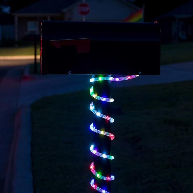 A black mailbox on a black post is wrapped with a colorful LED rope light that spirals downwards, emitting blue, green, red, and white light at night. The background shows a blurred residential street scene.