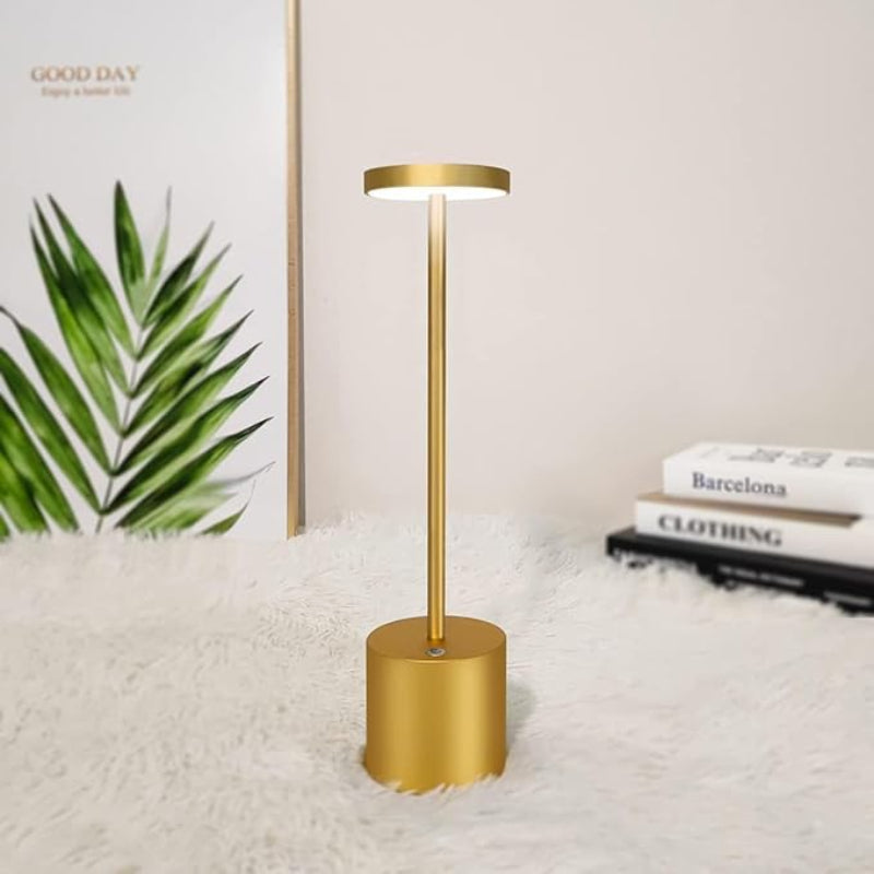 A cordless LED table lamp with a brushed gold finish, featuring a cylindrical base, a slender stem, and a circular light panel emitting a warm glow upwards, positioned on a fluffy white surface next to a framed print of palm leaves and a stack of books. The background is a plain light wall.