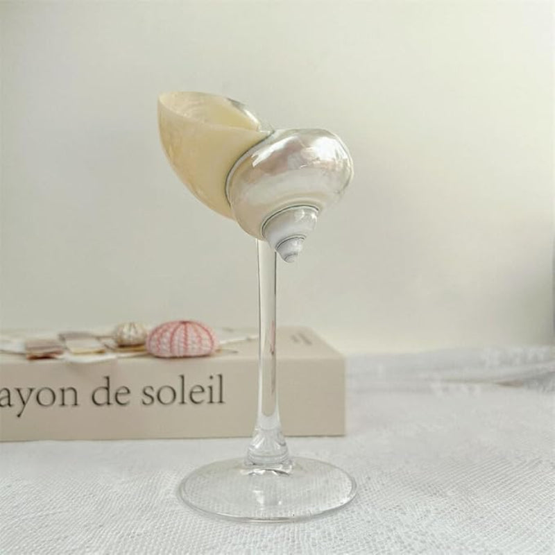 A Sharplace Conch Shell Wine Glass Cup Martini Glass with a clear stem and pearlescent seashell cup, displayed on a surface with a book titled "ayon de soleil" and two small sea urchin shells in the background.