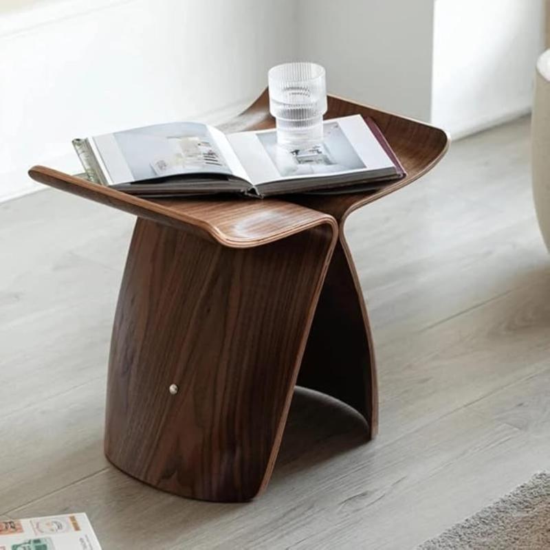 A warm-toned, eye-level close-up shows a Hans Wegner Butterfly Stool made of bent plywood. An open book and a clear drinking glass are placed on its curved top surface. The stool sits on a light-colored wooden floor. A portion of a magazine is visible in the lower left corner.