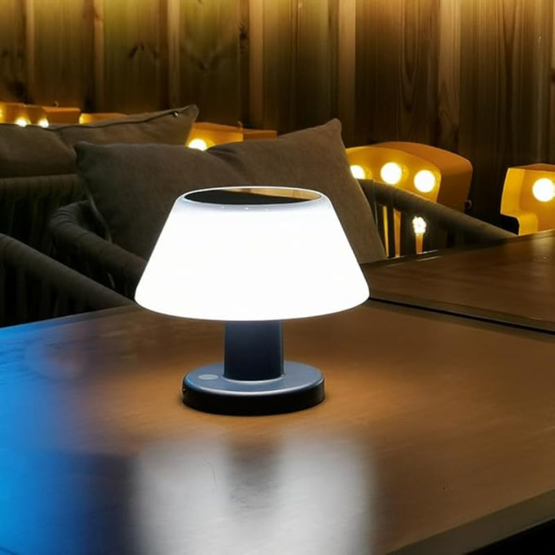 A small, solar-powered table lamp with a white, slightly tapered shade and a dark base illuminates a wooden table in a dimly lit indoor or outdoor setting. The lamp casts a soft glow on the tabletop. Blurred seating with pillows and warm string lights are visible in the background, suggesting a cozy ambiance.