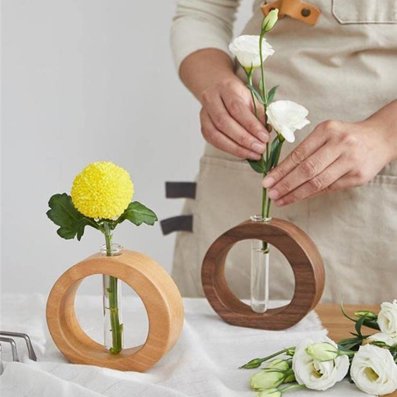 Two circular wooden vases, one light and one dark, each hold a clear glass test tube with flowers. A person's hands are arranging white flowers in the darker vase. A yellow chrysanthemum is in the lighter vase.