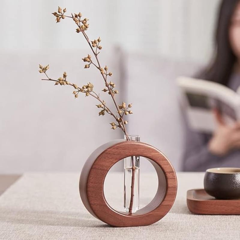 A dark wooden circular vase holds a clear glass test tube with a delicate branch bearing small buds. It sits on a textured gray surface. In the blurred background, a person appears to be reading.