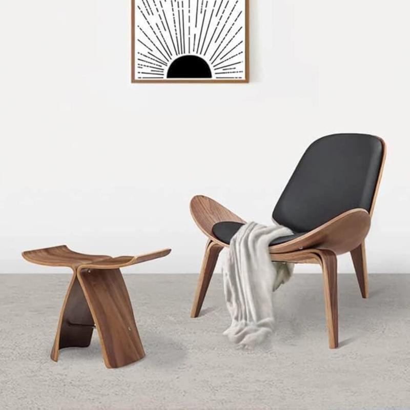 A modern living space features a sleek, dark-cushioned wooden chair draped with a light-colored throw, next to a smaller, matching wooden stool. A framed graphic print of a rising sun hangs on the white wall above. The floor is a light gray.