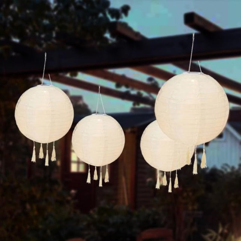 The image shows four round, cream-colored paper lanterns hanging against a backdrop of green foliage and a wooden structure. Each lantern has a series of tassels hanging from the bottom