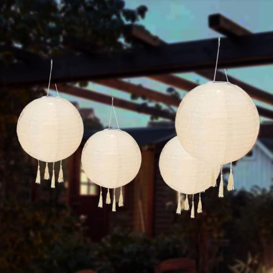 The image shows four round, cream-colored paper lanterns hanging against a backdrop of green foliage and a wooden structure. Each lantern has a series of tassels hanging from the bottom