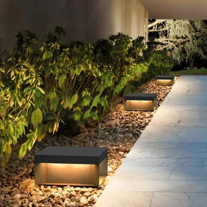 Three modern, rectangular outdoor lights with dark tops and glowing warm-toned panels, illuminating a stone pathway bordered by pebbles and lush greenery at night.