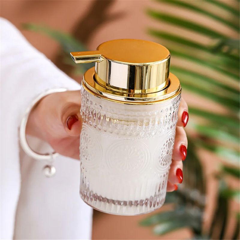 A hand with red nails holding a clear glass soap dispenser filled with white liquid. The dispenser has ornate patterns and a gold pump. A bracelet is visible on the wrist.