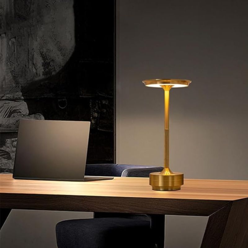 Sleek gold-colored table lamp with a modern, minimalist design. Features a slim, cylindrical stem and a flat, disc-shaped LED light source. Placed on a wooden desk beside a laptop in a dimly lit room.