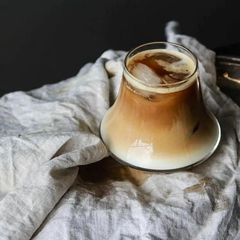 A uniquely shaped glass filled with iced coffee, showcasing a beautiful gradient of creamy milk blending with dark coffee, placed on a soft linen cloth.