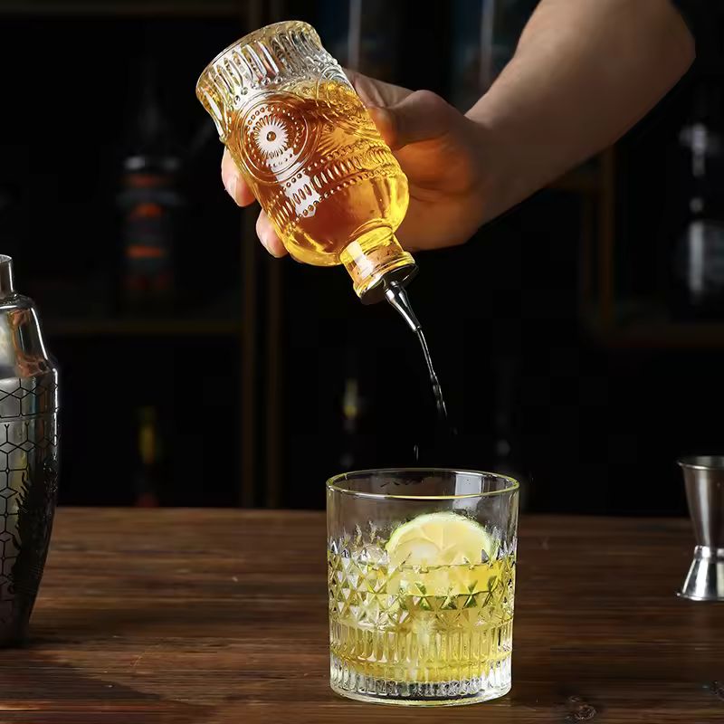 A hand pours a golden liquid from an ornate glass bitters bottle with a metal spout into a cocktail with ice and lemon slices.