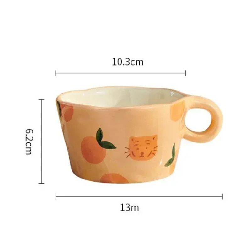 A close-up shot of a peach-colored ceramic mug with hand-painted peach illustrations and a small, stylized cartoon tiger face. Measurement lines and text indicate the following dimensions: 10.3 centimeters width, 6.2 centimeters height, and 13 meters overall width including the handle. The mug has a rounded handle and a slightly uneven rim, showcasing its handcrafted nature. The focus is on the mug's design and its dimensions.