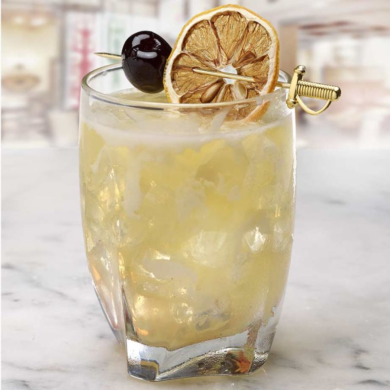 A yellow cocktail in a faceted glass, garnished with a dried lemon slice, cherry, and sword-shaped gold cocktail pick.