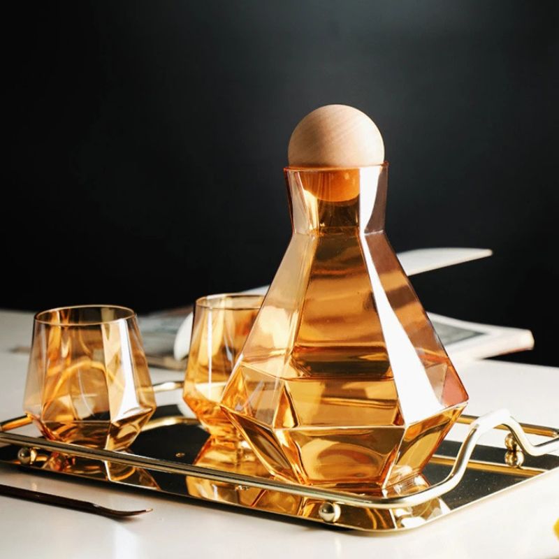 Amber glass pitcher and two matching faceted glasses on a gold tray. A wooden ball lid is on the pitcher. The background is dark with a white surface visible.