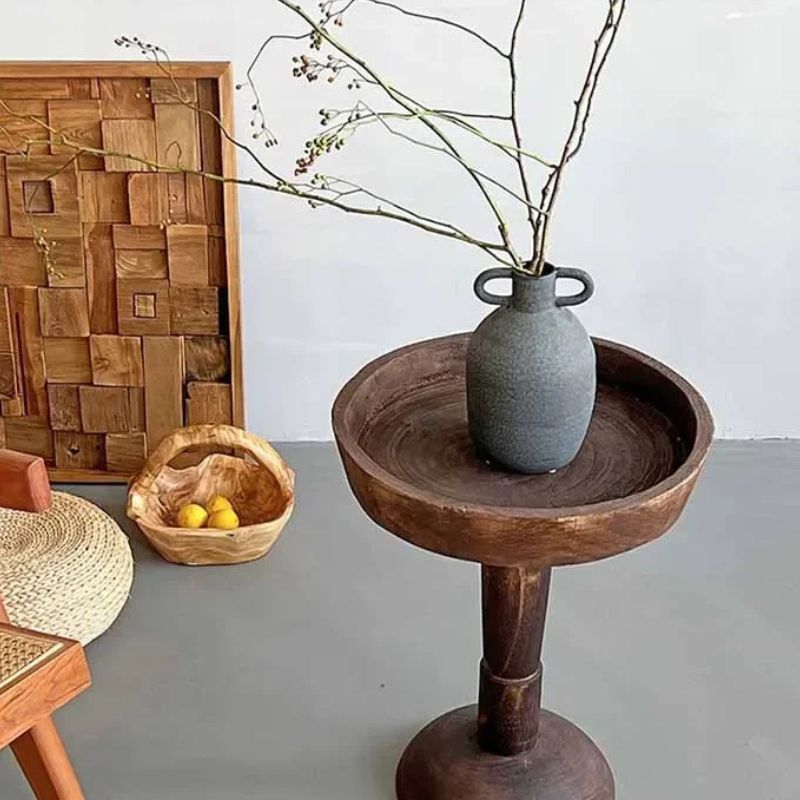 A rustic interior scene with a dark wood, round side table holding a gray vase with bare branches, a wooden basket with lemons, a patterned wooden wall hanging, and a woven stool.