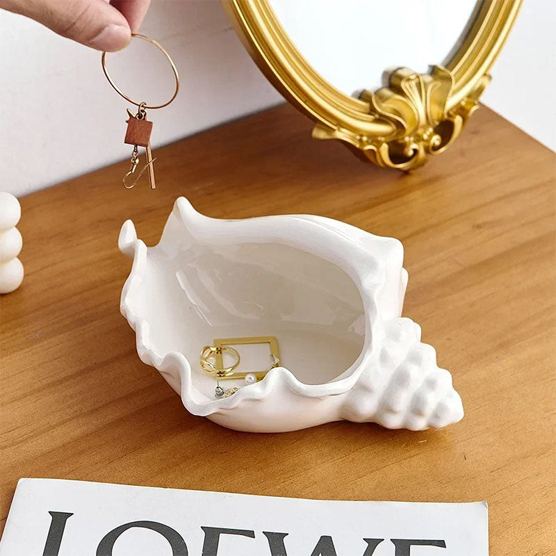 A white ceramic seashell-shaped jewelry dish on a wooden surface, holding gold earrings, with a gold-framed mirror, a hand holding a hoop earring, and a magazine nearby.